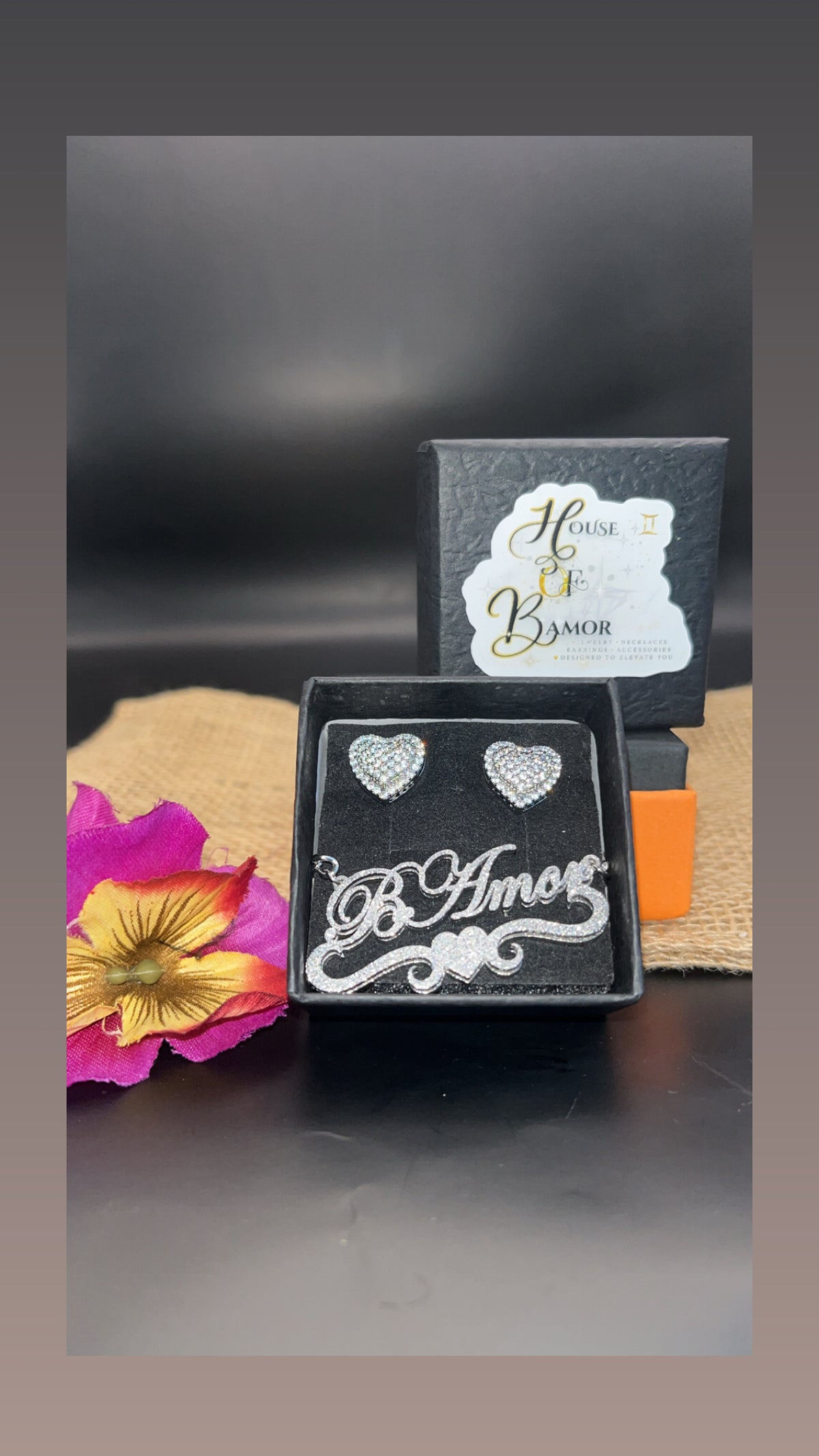 A pair of silver heart stud earrings displayed in a black gift box with a decorative 'B Amor' script plaque, alongside the brand box 'House of Amor' and a pink flower prop on a dark surface.