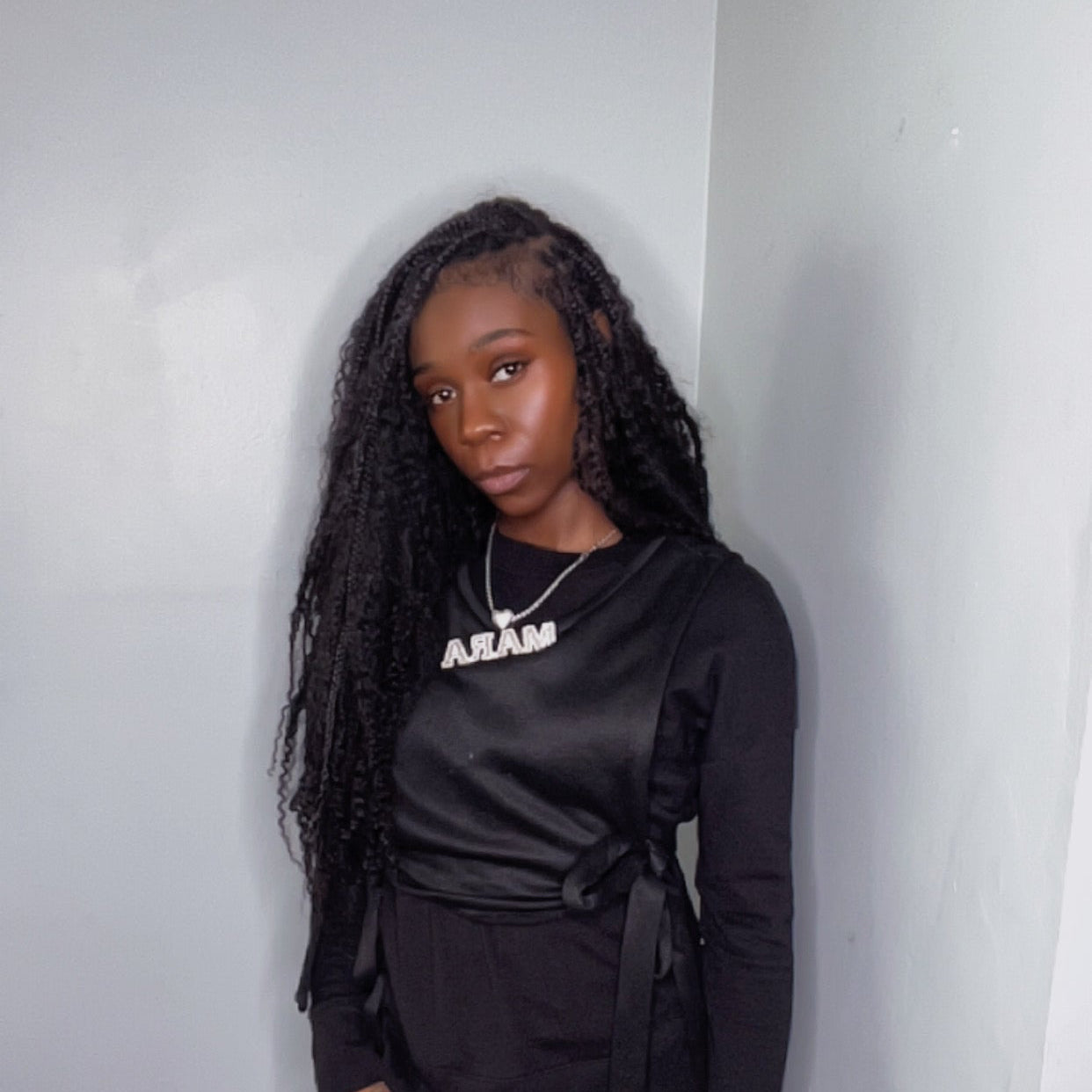 Person wearing a black outfit with a logo, standing against a plain wall. model is wearing european human kinky braid hair 
