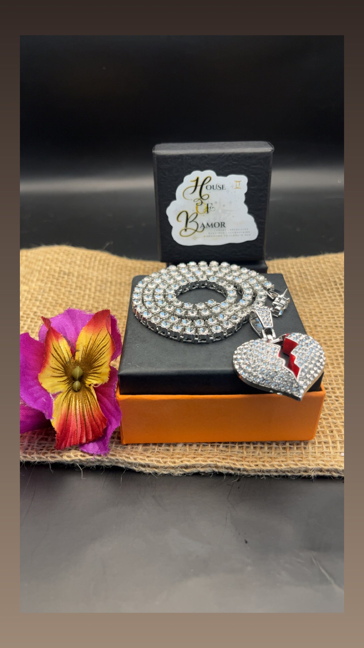 A silver-toned necklace with a heart-shaped pendant, displayed on top of an orange and black jewelry box. The necklace is covered in clear rhinestones, and the pendant has a red heart-shaped accent.