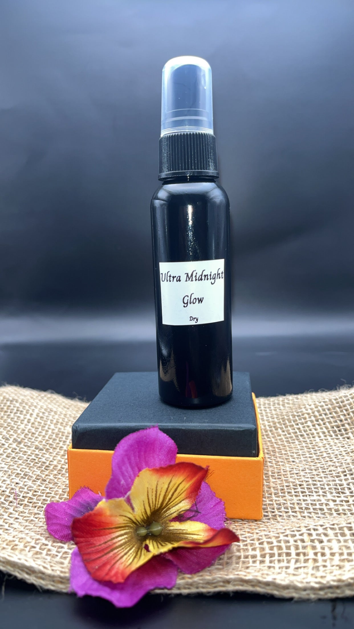 Bottle of 'Ultra Midnight Glow' on an orange stand with a colorful flower in front against a dark background