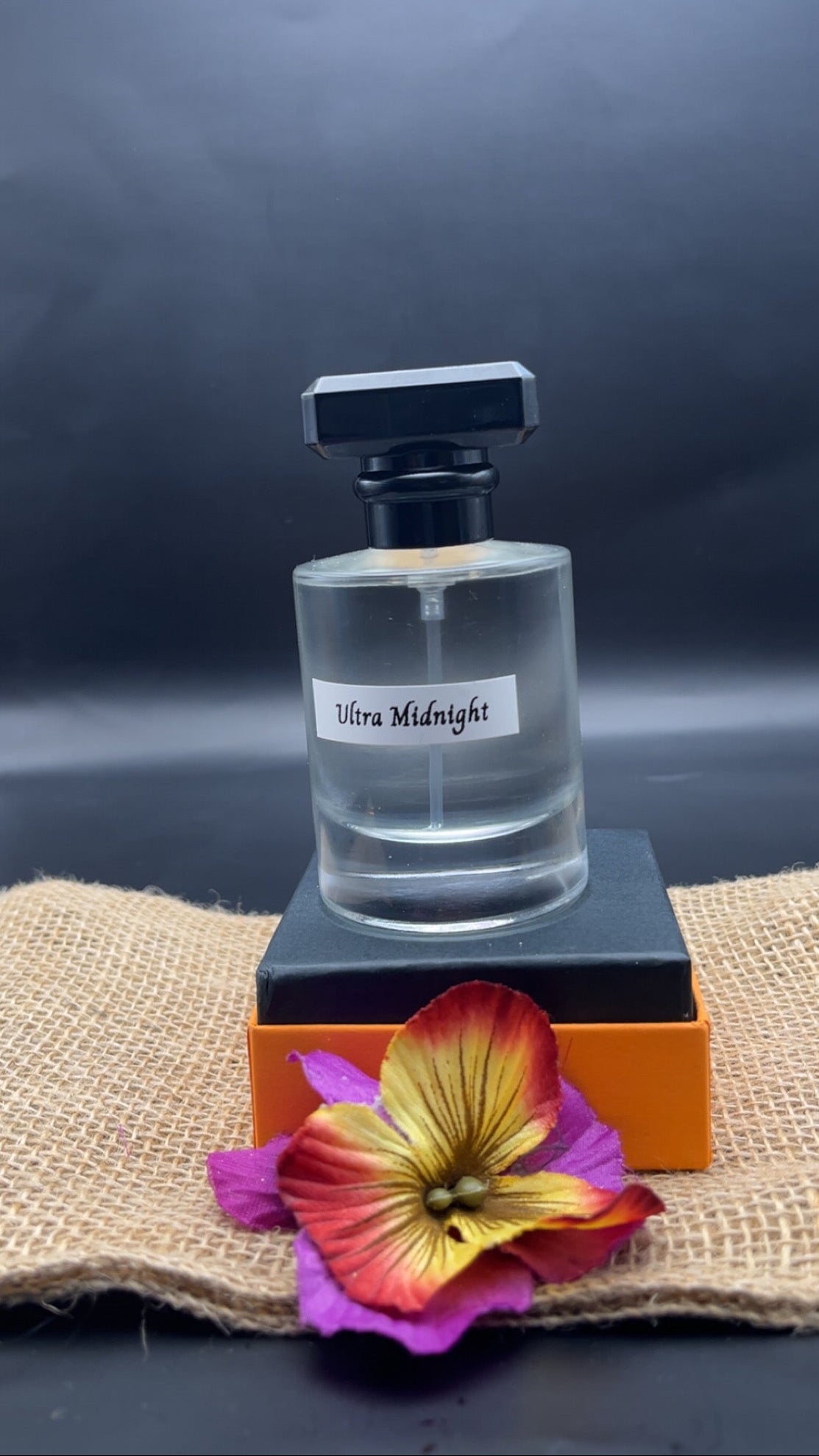 Perfume bottle labeled 'Ultra Midnight' on an orange stand with a colorful flower on a dark background