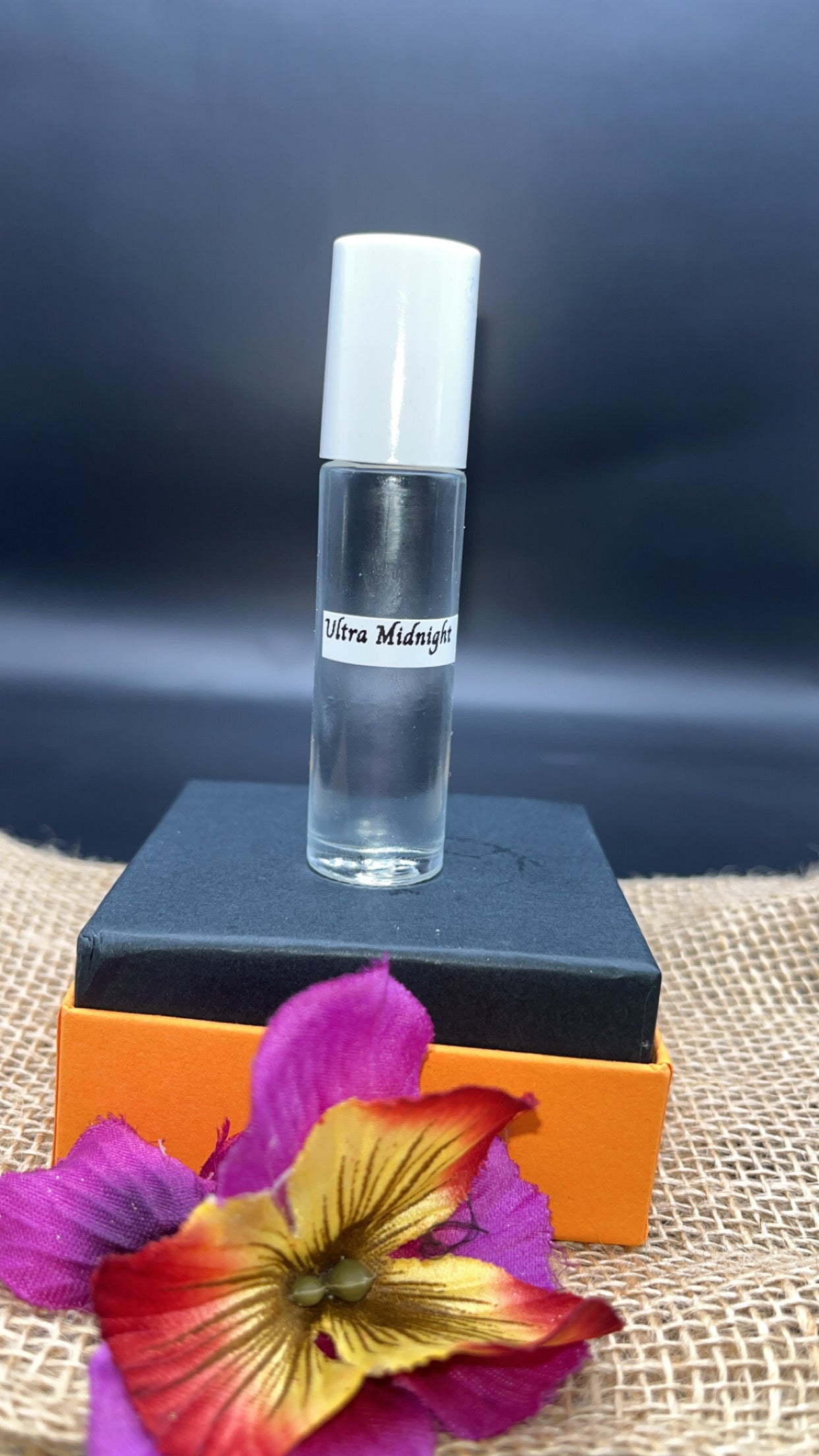 Small clear bottle with a label on an orange stand, with a dark background and a colorful flower in the foreground.